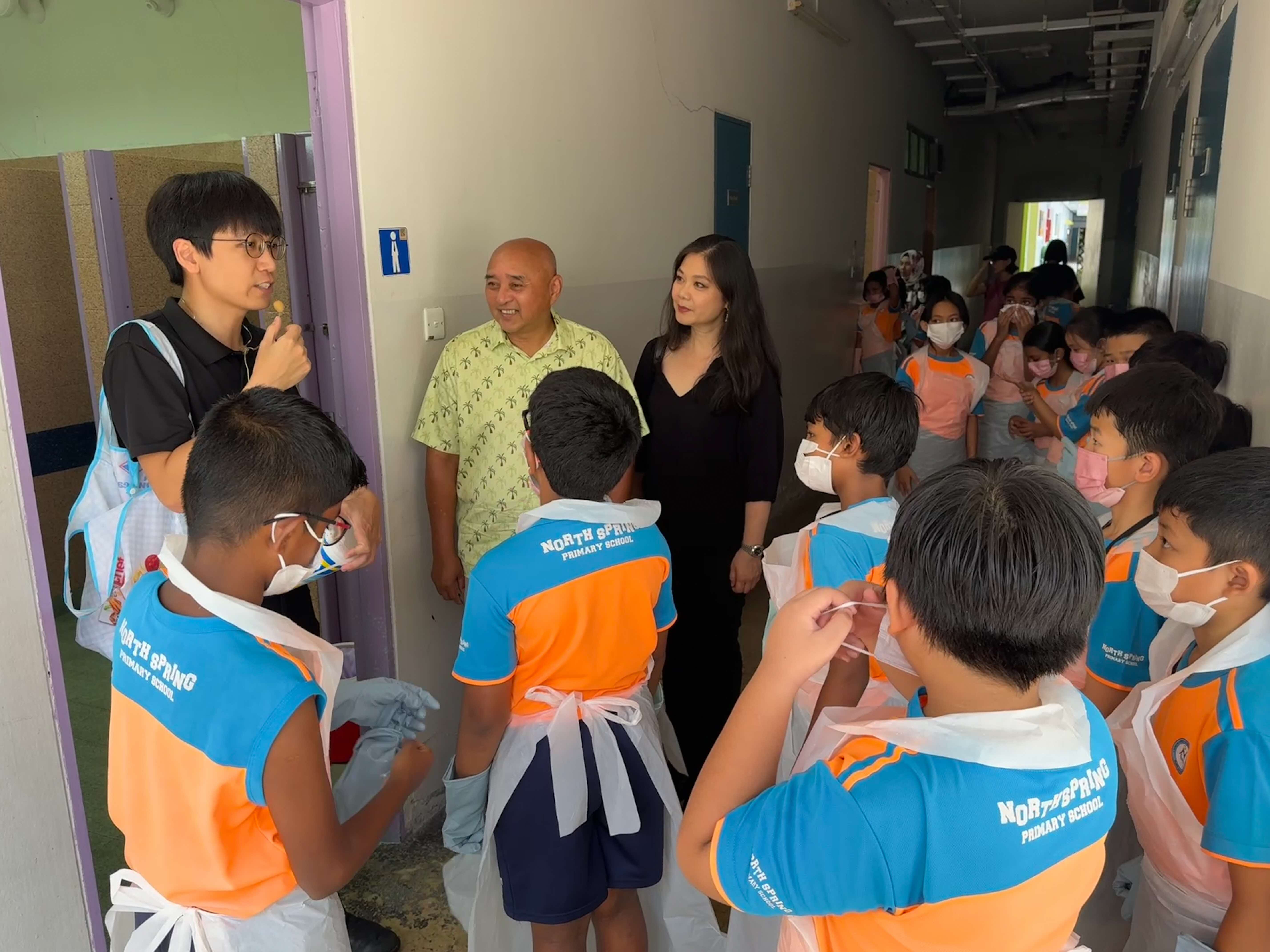 A group of North Spring Primary School students in orange and blue uniforms putting on aprons and masks in a school corridor, preparing for a cleaning activity. Two adults and a facilitator stand nearby, engaging with the students. More students can be seen lining up in the background.